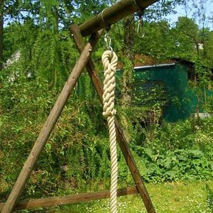 Puede incluir: Un columpio de madera con una gruesa cuerda blanquecina colgando de un gancho de metal. La cuerda está anudada en la parte superior. El columpio está en una zona de césped con árboles al fondo.