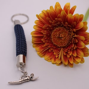 Könnte beinhalten: Ein blauer Seil-Schlüsselanhänger mit einem silbernen Schlüsselring und einem silbernen Schwimmer-Anhänger. Der Schlüsselanhänger liegt neben einer leuchtend orangefarbenen und gelben Gerbera.