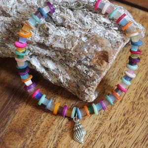 Könnte beinhalten: Ein farbenfrohes Armband mit kleinen, flachen, runden Perlen in Rosa, Orange, Gelb, Grün, Blau und Lila. Ein kleiner silberner Muschel-Anhänger hängt am Armband. Das Armband wird auf einem Stein präsentiert.