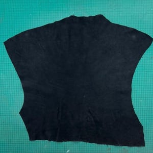 Puede incluir: Un trozo de cuero negro, de forma irregular, posiblemente para artesanía o trabajos en cuero. El cuero tiene un acabado mate y parece estar cortado de una piel más grande. Los bordes son crudos y sin terminar.