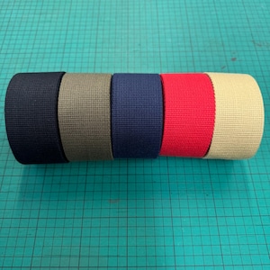 Puede incluir: Un rollo de cincha de colores en varios tonos: negro, verde oliva, azul marino, rojo y beige. La cincha está tejida apretadamente y parece duradera, adecuada para diversas aplicaciones artesanales e industriales. Los colores son vibrantes y están distribuidos uniformemente.