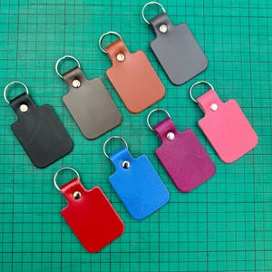 May include: A collection of rectangular leather keychains in various colors, including black, gray, brown, red, blue, purple, and pink. Each keychain has a silver metal ring and a rivet securing the leather tab.