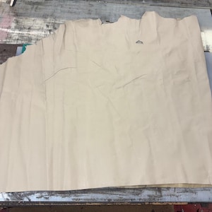May include: A large, irregularly shaped piece of light beige leather. The leather has a soft, matte finish and visible creases and imperfections. The edges are uneven, suggesting it's been cut from a larger hide. The material appears to be natural.