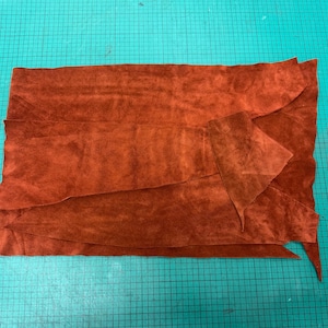 Può includere: Una collezione di pezzi di pelle scamosciata color ruggine, disposti su un tappetino da taglio verde. La pelle ha un aspetto morbido e strutturato, con pieghe e bordi visibili. I pezzi variano per dimensioni e forma, suggerendo che siano tagliati per un progetto artigianale.