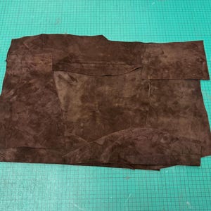 Op de afbeelding: Een verzameling donkerbruine leren stukken, mogelijk voor knutselen. Het leer heeft een gevlekt uiterlijk met kleurvariaties, wat wijst op een natuurlijke afwerking. De stukken zijn in verschillende vormen gesneden, waaronder een zakvorm.