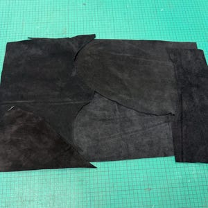 May include: Various pieces of black suede leather are laid out on a green cutting mat. The leather pieces are of different sizes and shapes, including some with curved edges and some rectangular.