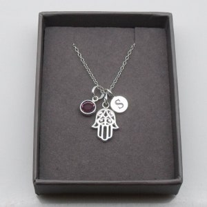 May include: A silver necklace featuring a hamsa hand charm, a small purple gemstone, and a silver charm engraved with the letter 'S'. The necklace is presented in a grey box.