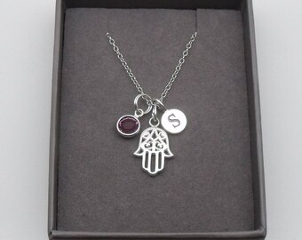 May include: A silver necklace with a hamsa hand charm, a small purple gemstone, and a silver charm with the letter 'S' engraved on it. The necklace is displayed in a gray box.
