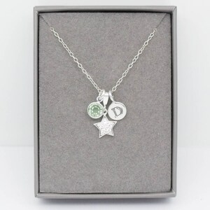 May include: A silver necklace with three charms: a round green gemstone, a small disc with the letter "D", and a star-shaped charm encrusted with small, clear crystals. The necklace is displayed in a gray jewelry box.