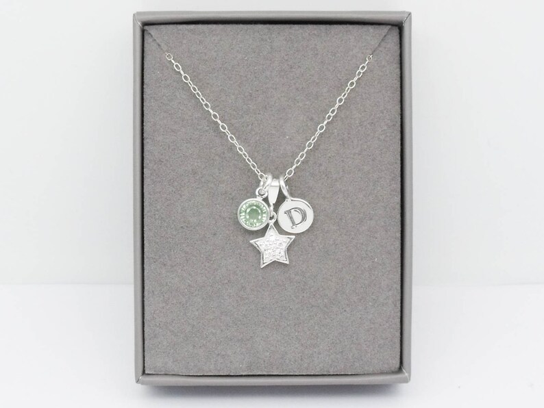 May include: A silver necklace with three charms: a round green gemstone, a small disc with the letter "D", and a star-shaped charm encrusted with small, clear crystals. The necklace is displayed in a gray jewelry box.