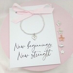 May include: A silver beaded bracelet with a heart charm is presented on a white card with the words "New beginnings. New strength." A pink ribbon and several small charms are also visible. The background is a soft pink.