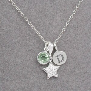 May include: A silver necklace with multiple charms. The necklace features a round green gemstone, a star-shaped charm with small clear stones, and a circular charm with the letter "D". The chain is delicate and the charms are suspended from it.