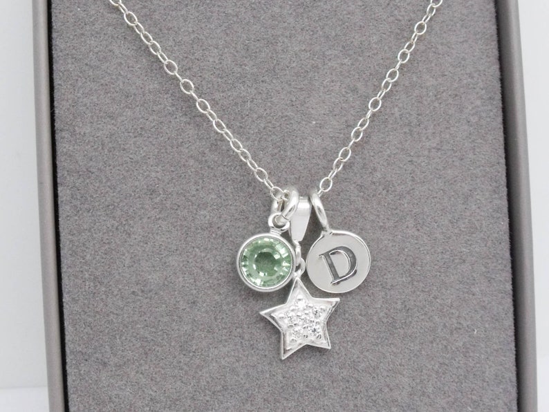 May include: A silver necklace with multiple charms. The necklace features a round green gemstone, a star-shaped charm with small clear stones, and a circular charm with the letter "D". The chain is delicate and the charms are suspended from it.