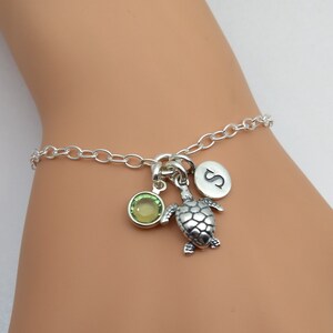 May include: A silver charm bracelet with a turtle charm, a green gemstone, and a circular charm with the letter "S". The bracelet is on a wrist.