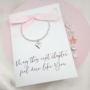 May include: A beaded bracelet with a heart charm is displayed on a white card with a pink ribbon. The card has the text "May this next chapter feel more like You". Several charms in heart and star shapes are also shown.
