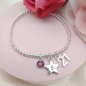 May include: A silver beaded bracelet with a star charm, a crystal charm, and a charm with the number 21. The star charm has the letter S engraved on it.