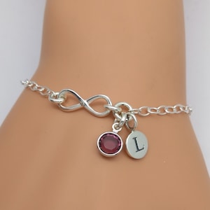 May include: Silver infinity charm bracelet with a purple crystal and a charm with the letter L.