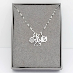 May include: Silver necklace with a paw print charm, a small round charm with a clear stone, and a round charm with the letter 'S' engraved on it.
