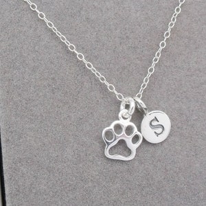 May include: A silver-colored necklace with a paw print charm and a circular charm with the letter "S". The necklace is displayed on a gray surface.