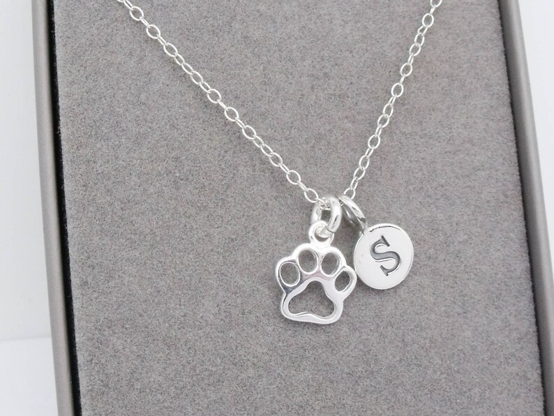May include: A silver-colored necklace with a paw print charm and a circular charm with the letter "S". The necklace is displayed on a gray surface.