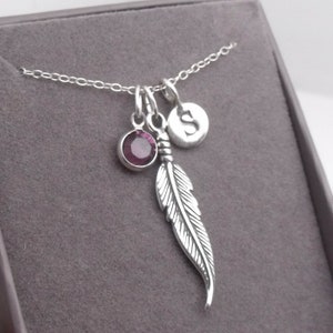 May include: Silver-toned necklace with a feather pendant, a purple gemstone, and a circular charm with the letter "S". The chain and charms are displayed against a gray background.