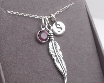 May include: Silver-toned necklace with a feather pendant, a purple gemstone, and a circular charm with the letter "S". The chain and charms are displayed against a gray background.