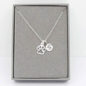 May include: A silver necklace with a paw print charm and a round charm with the letter "S" inside a gray jewelry box. The necklace chain is delicate and the charms are small.