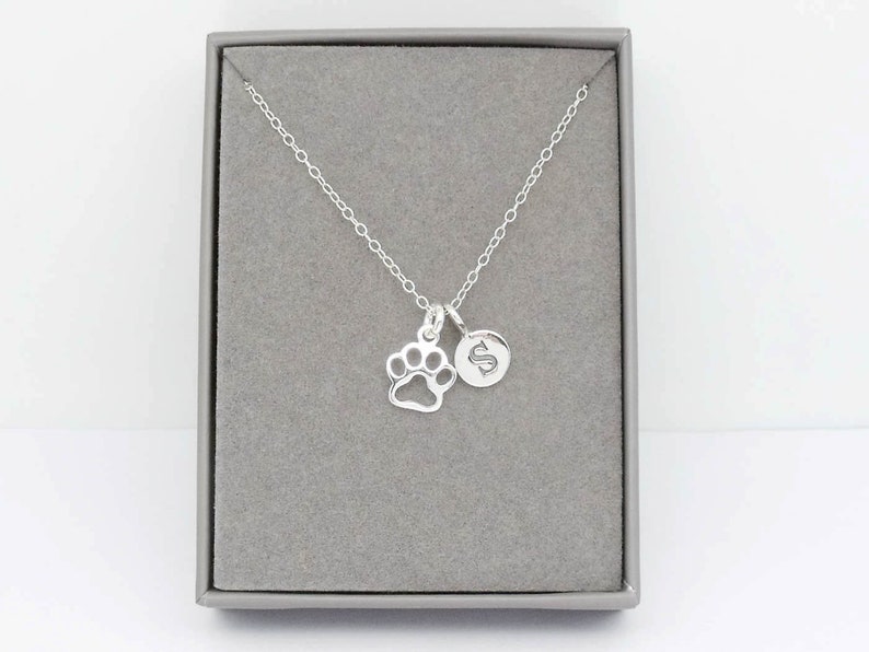 May include: A silver necklace with a paw print charm and a round charm with the letter "S" inside a gray jewelry box. The necklace chain is delicate and the charms are small.