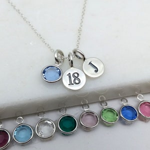 May include: A silver necklace with three pendants: a light blue gemstone, a disc with the number 18, and a disc with the letter "J". Additional gemstone pendants in various colors are visible below.