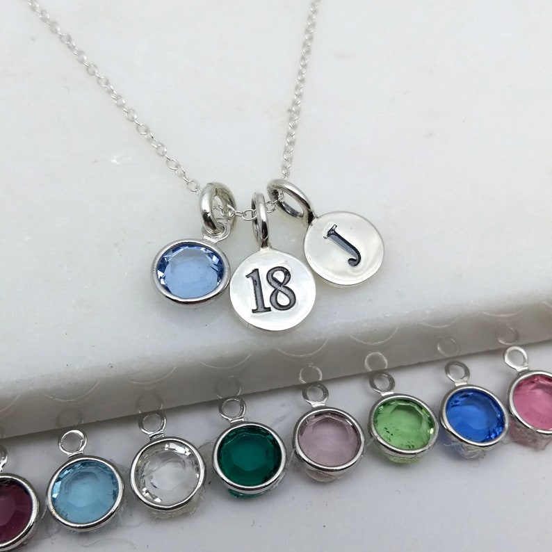May include: A silver necklace with three pendants: a light blue gemstone, a disc with the number 18, and a disc with the letter "J". Additional gemstone pendants in various colors are visible below.