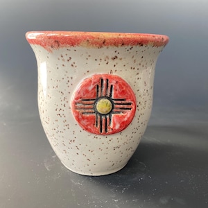 May include: A white ceramic mug with a red and yellow symbol on the front. The mug has a speckled glaze.