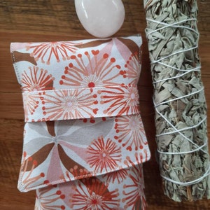 May include: A white fabric pouch with a floral pattern in shades of pink and orange. The pouch is folded over and has a pink rose quartz crystal and a bundle of white sage smudge stick next to it.