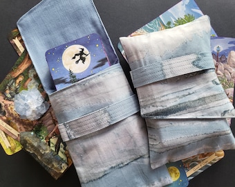 Op de afbeelding: Twee blauwe en grijze stoffen zakjes met een strik sluiting. Het zakje aan de linkerkant heeft een zakje aan de voorkant met een kaart erin. Het zakje aan de rechterkant is gesloten. Beide zakjes hebben een aquarel-achtig ontwerp.