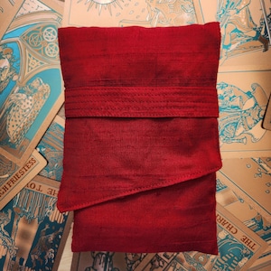 Puede incluir: Una bolsa roja para cartas del tarot, de forma rectangular, con una banda texturizada. La bolsa está rodeada de cartas del tarot con ilustraciones azules, doradas y marrones. La bolsa está hecha de una tela lisa y brillante.