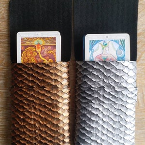 Op de afbeelding: Twee tarotkaarthouders met een zwarte flap en een schubbenpatroon. De houder aan de linkerkant is koperkleurig en de houder aan de rechterkant is zilverkleurig. De houder aan de linkerkant heeft een tarotkaart met het nummer XI. De houder aan de rechterkant heeft een tarotkaart met het nummer III.