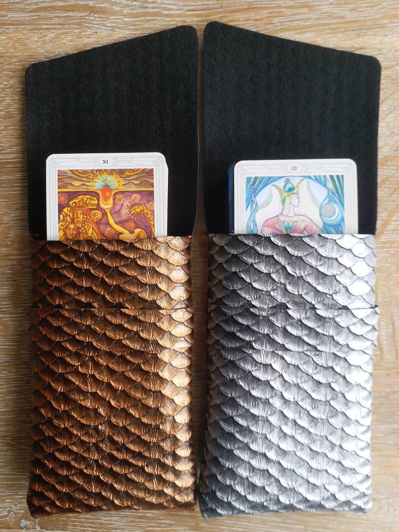 Op de afbeelding: Twee tarotkaarthouders met een zwarte flap en een schubbenpatroon. De houder aan de linkerkant is koperkleurig en de houder aan de rechterkant is zilverkleurig. De houder aan de linkerkant heeft een tarotkaart met het nummer XI. De houder aan de rechterkant heeft een tarotkaart met het nummer III.