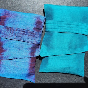 Puede incluir: Dos bolsos de mano de seda. Uno es turquesa, el otro azul con detalles morados. Ambos tienen un cierre de solapa asegurado por una banda de tela a juego. Los bolsos son rectangulares y parecen hechos a mano.