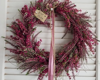May include: A circular wreath made of vibrant magenta heather flowers with green stems. A small beige rectangular tag with the handwritten text "Herbst Zauber" and a heart symbol is attached. A long pink ribbon and twine hang from the top center.