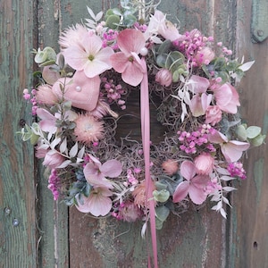 Könnte beinhalten: Ein kreisförmiger Blumenkranz mit einer Vielzahl von rosa Blüten, darunter Hortensien und kleinere Blüten, mit grünen Blättern. Ein rosa Band und eine kleine herzförmige Dekoration sind enthalten. Der Kranz wird an einer Schnur aufgehängt.