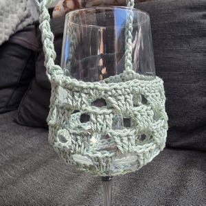 May include: A clear glass wine goblet encased in a light sage green crocheted sleeve. The sleeve features an openwork square pattern and includes a matching crocheted handle attached to the sides, wrapping around the bowl of the glass.