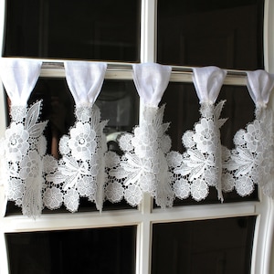 Peut inclure: Une cantonnière de fenêtre en dentelle blanche avec des motifs floraux et de feuilles. La cantonnière est suspendue à une tringle argentée, avec du tissu blanc en haut. La dentelle détaillée ajoute une touche délicate à la fenêtre.