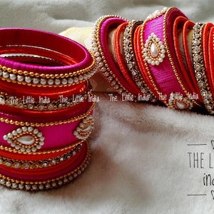 Könnte beinhalten: Eine Sammlung farbenfroher Armreifen in Pink, Orange und Rot, verziert mit Perlen und goldfarbenen Akzenten. Die Armreifen sind gestapelt und auf einer weichen, weißen Oberfläche angeordnet. Der Text "THE LITTLE india" ist sichtbar.