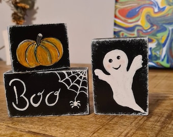 May include: Three black wooden blocks with Halloween decorations. The top block has a yellow pumpkin, the middle block has a spider web and the word "Boo" and the bottom block has a white ghost.