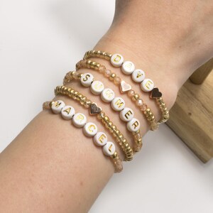 Könnte beinhalten: Goldene Armbänder mit weißen Buchstabenperlen, die Namen und das Wort „SUMMER“ bilden. Jedes Armband hat ein anderes Design mit herzförmigen Anhängern und transparenten Perlen. Die Armbänder werden am Handgelenk getragen.