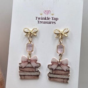 May include: Gold-toned dangle earrings featuring a bow and a stack of books design. Each earring has a gold bow at the top, a pink rectangular gem, and a stack of books with a pink bow. The text "Twinkle Tap Treasures" is visible.