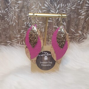 May include: A pair of dangle earrings with a pink leather base and gold glitter accents. The earrings are displayed on a stand with the text "Twinkle Tap Treasures".