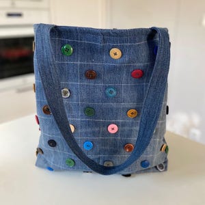 Może przedstawiać: Dżinsowa torba typu tote z wzorem kratki, ozdobiona różnokolorowymi guzikami. Torba posiada pasujący dżinsowy pasek. Guziki są w różnych kolorach, co dodaje zabawnego akcentu do projektu torby.