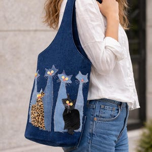 Op de afbeelding: Een denim draagtas met een grillig kattenontwerp. De tas heeft vijf geappliqueerde katten in verschillende kleuren en patronen, waaronder een luipaardprint kat. De katten hebben knoopogen en staan op een blauwe denim achtergrond.