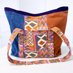 Può includere: Borsa tote patchwork con un pannello in denim, un pannello marrone e un pannello fantasia con tasca. La borsa ha un bordo viola e una tracolla fantasia. Il motivo include forme geometriche arancioni, rosa e viola.