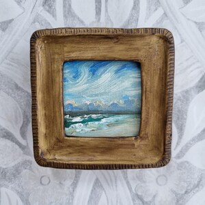 May include: A small, square oil painting of a seascape, framed in a rustic, textured, golden-brown frame. The artwork depicts a dynamic sky with swirling blue and white clouds above a turquoise ocean with white-capped waves. The painting is a miniature landscape.